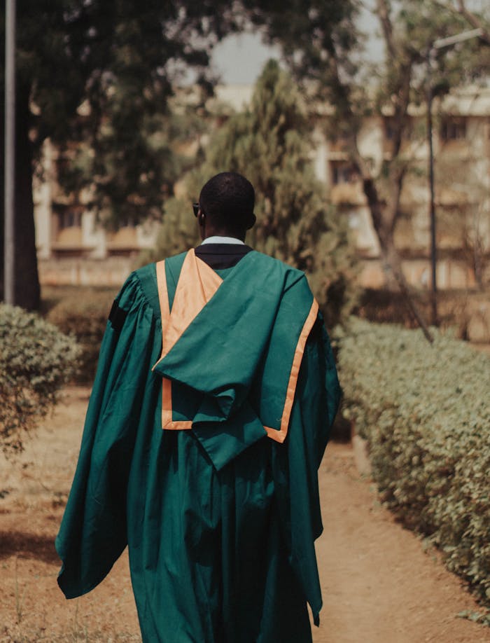 A graduate in green and orange gown walking outdoors, from behind.